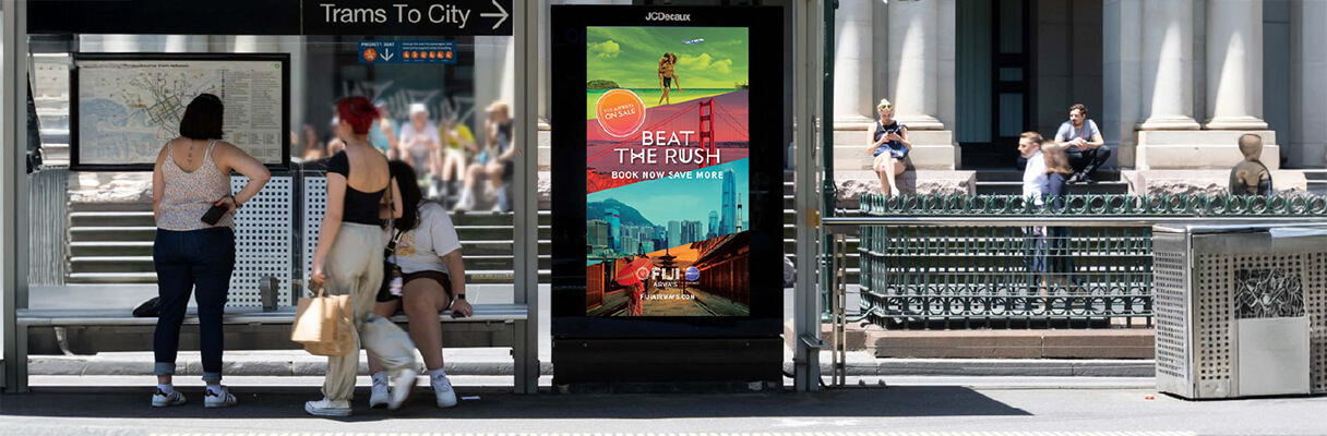 People waiting at a tram stop beside a digital billboard displaying a travel advertisement.